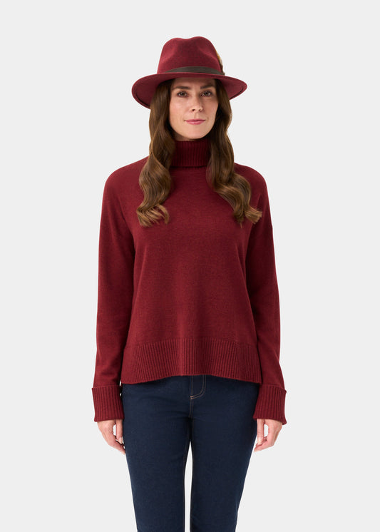 Woman in Alexa Ladies Wool and Cashmere Roll Neck in Berry, red hat, and blue jeans stands against a plain background.