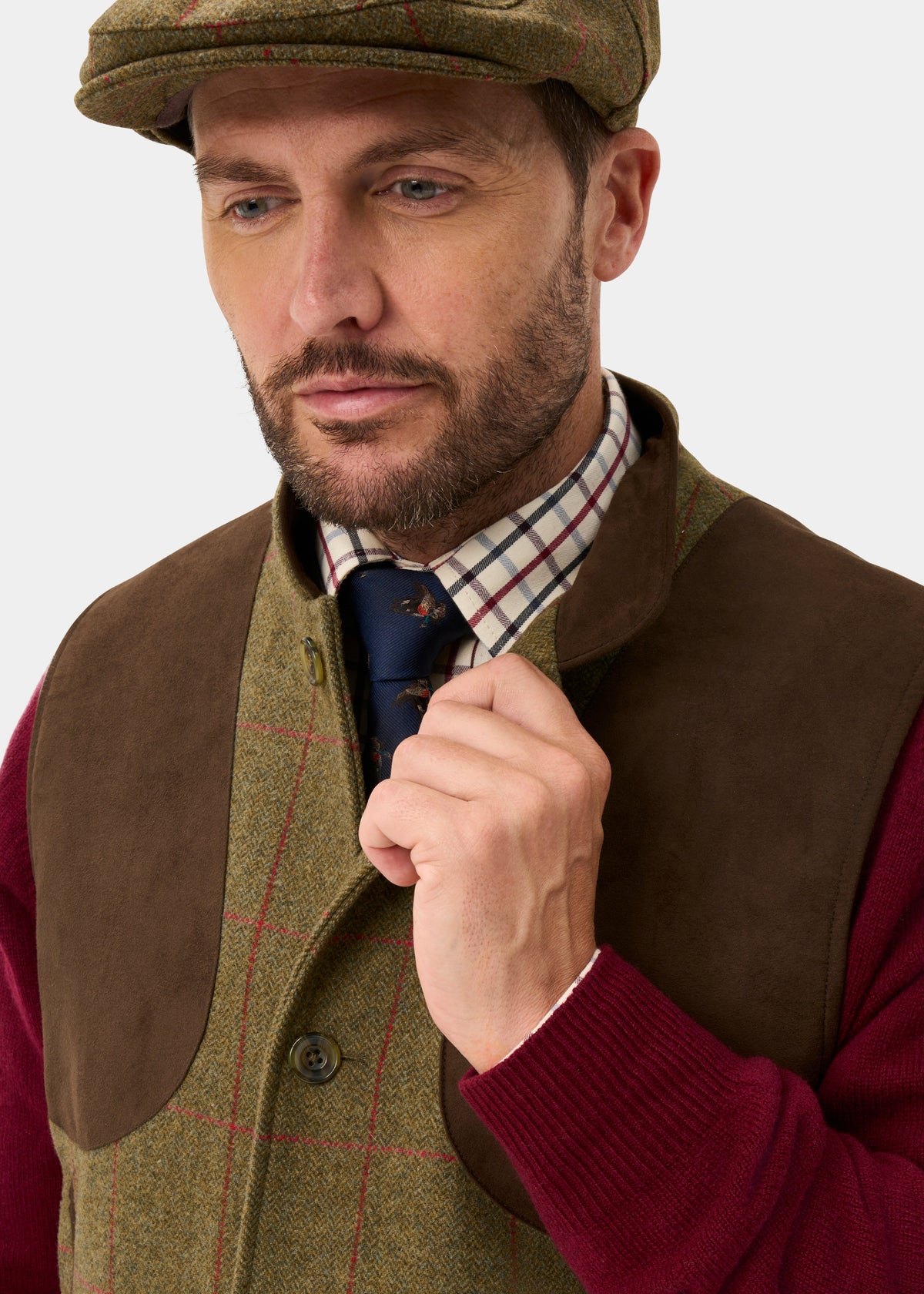 Man in a Combrook Sage Tweed Gilet, brown suede trim, checked shirt, tie, and cap adjusts his tie.