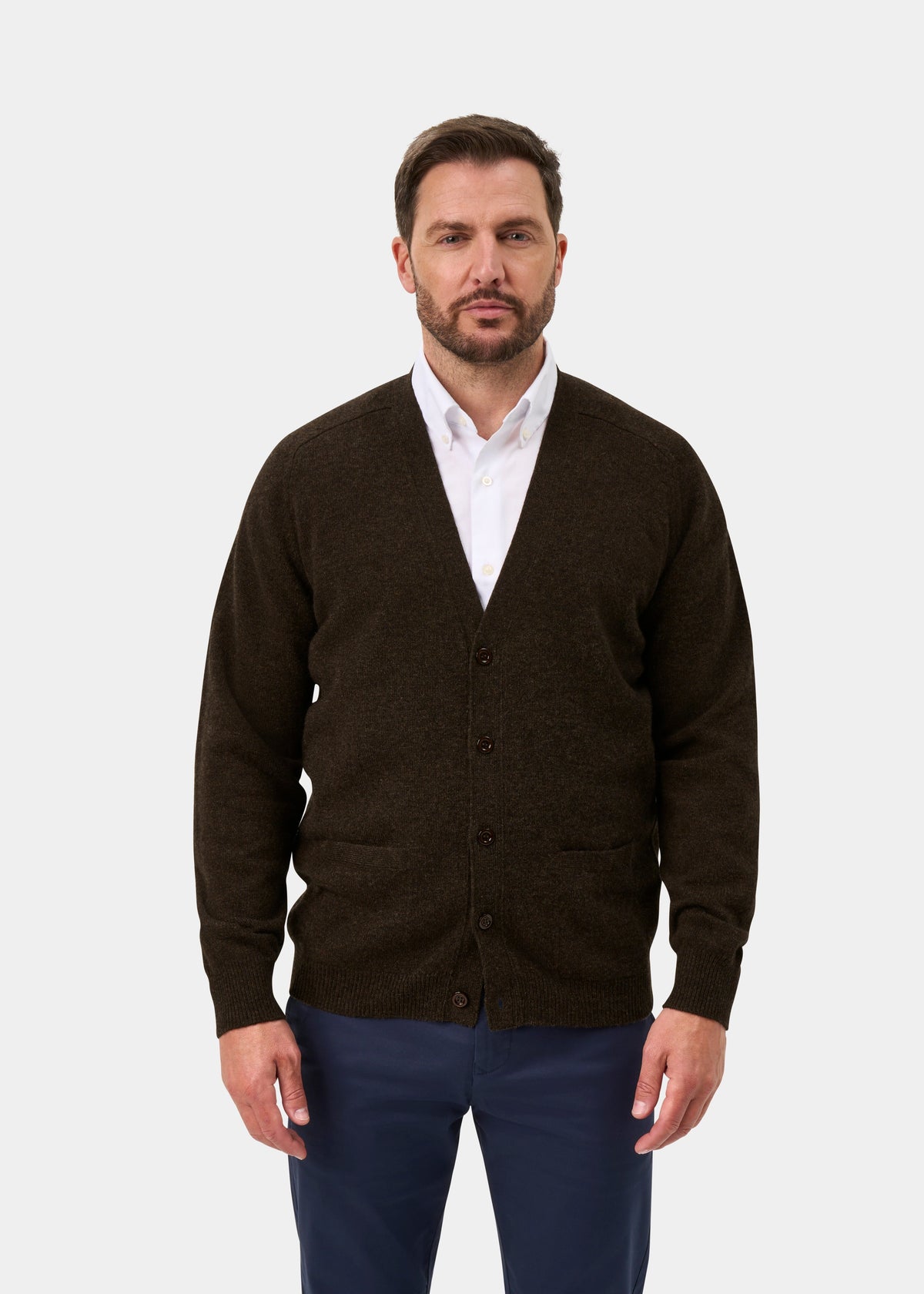 Man in Cornwall Lambswool V-Neck Cardigan in Cocoa, white shirt, navy pants, standing against a plain background.