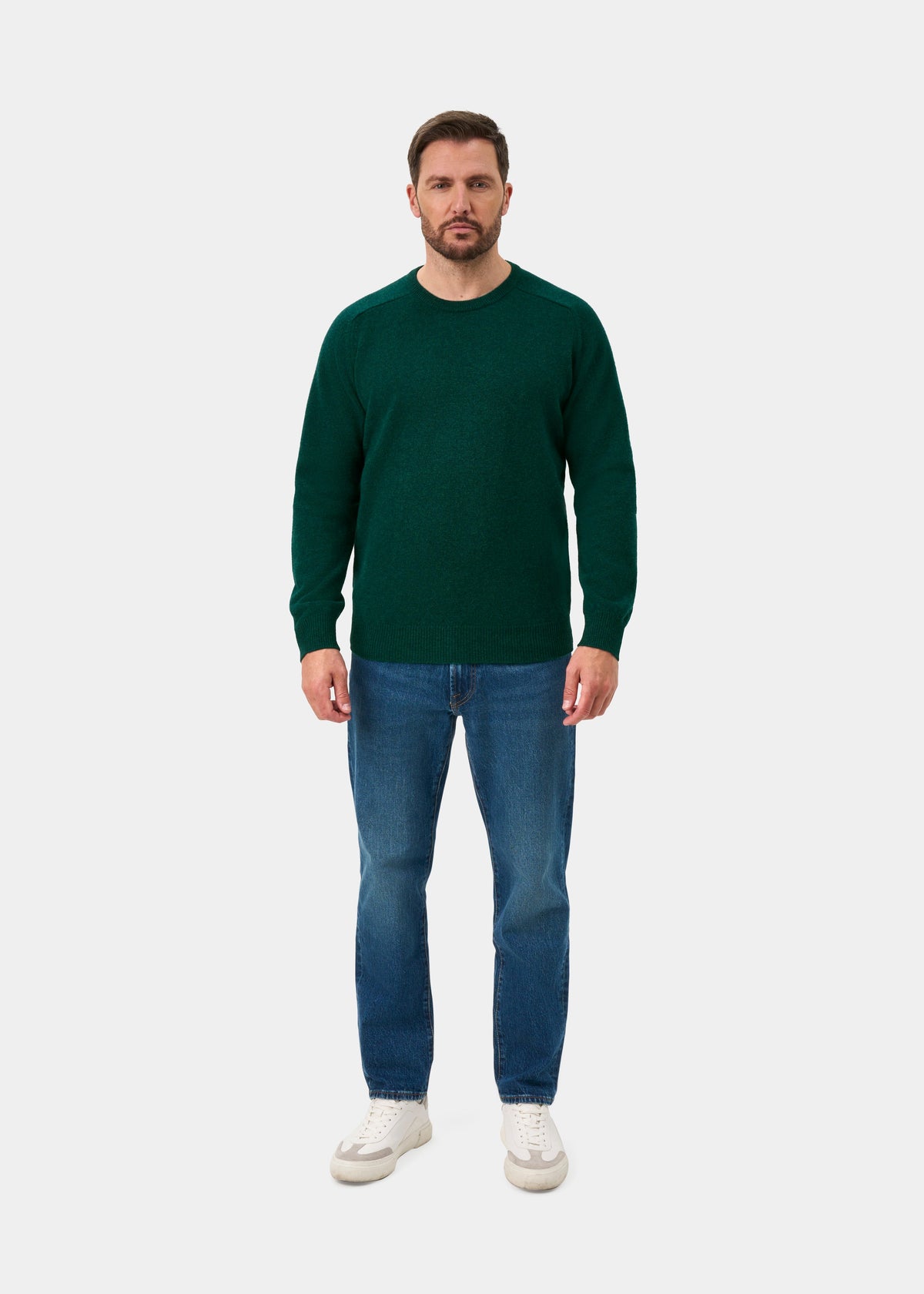 Man wears Dorset Lambswool Crew Neck Jumper in Mallard, blue jeans, and white sneakers against a plain white background.