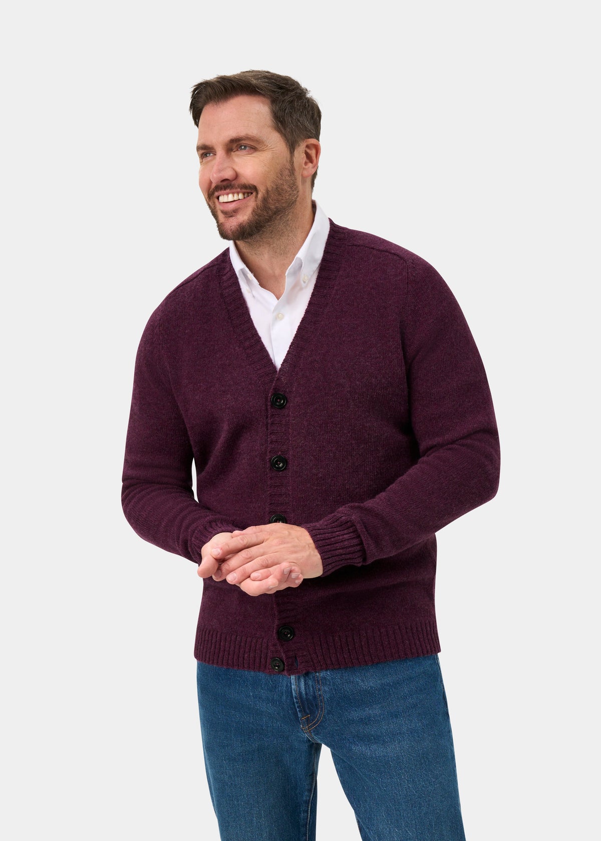 Smiling man in Kinloch Supersoft Shetland Wool V-Neck Cardigan in Elderberry, white shirt, blue jeans on gray background.