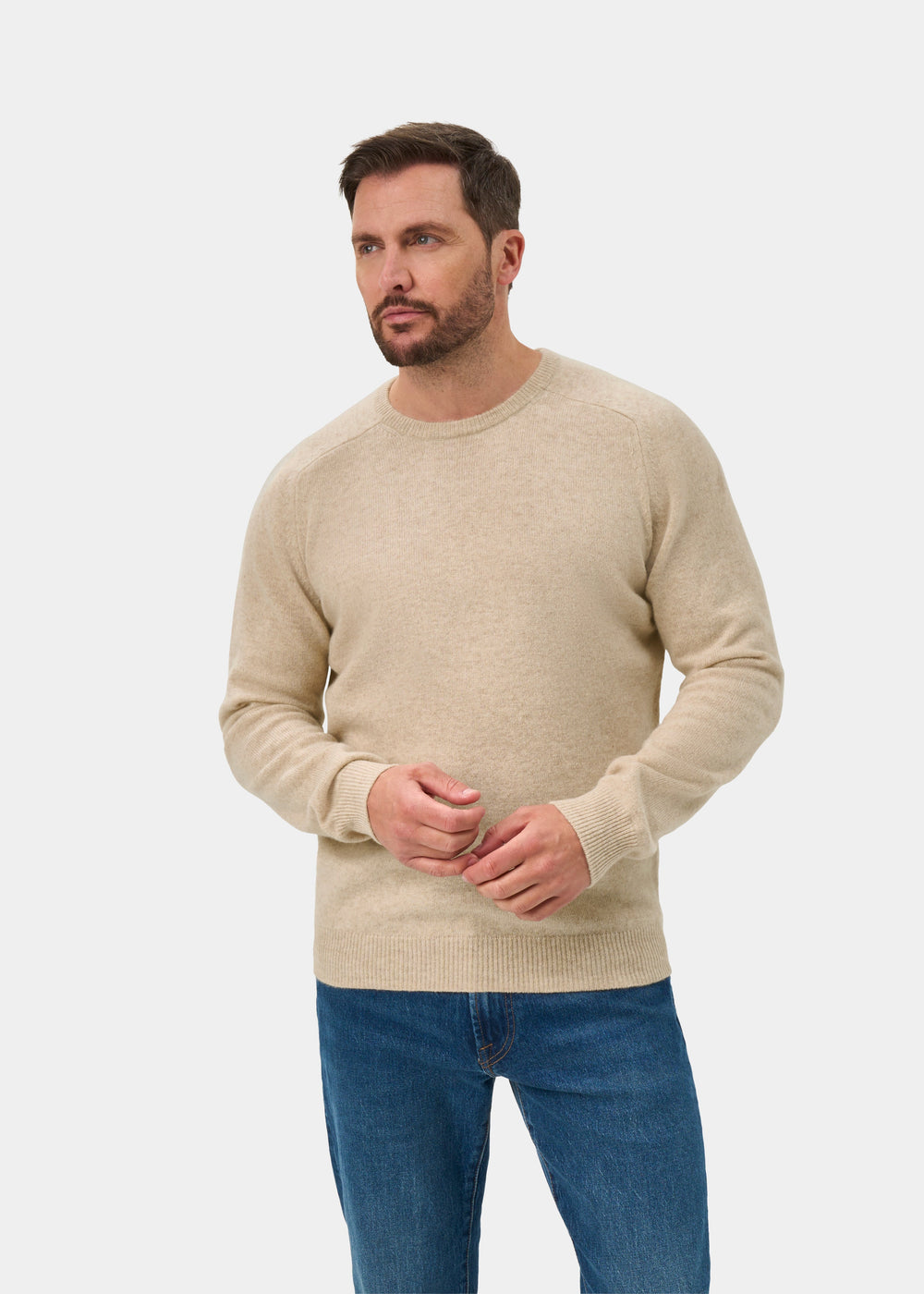 Man in a Lenzie Lambswool Crew Neck Jumper in Linen, regular fit, and blue jeans stands against a white backdrop.