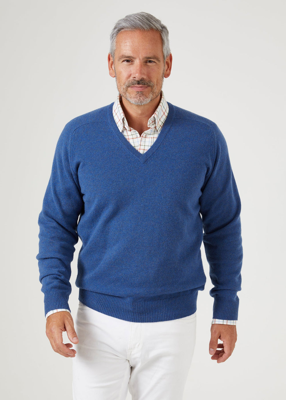 A man wearing a Selkirk Pure Cashmere V-Neck Jumper In Denim over a plaid collared shirt and white trousers.