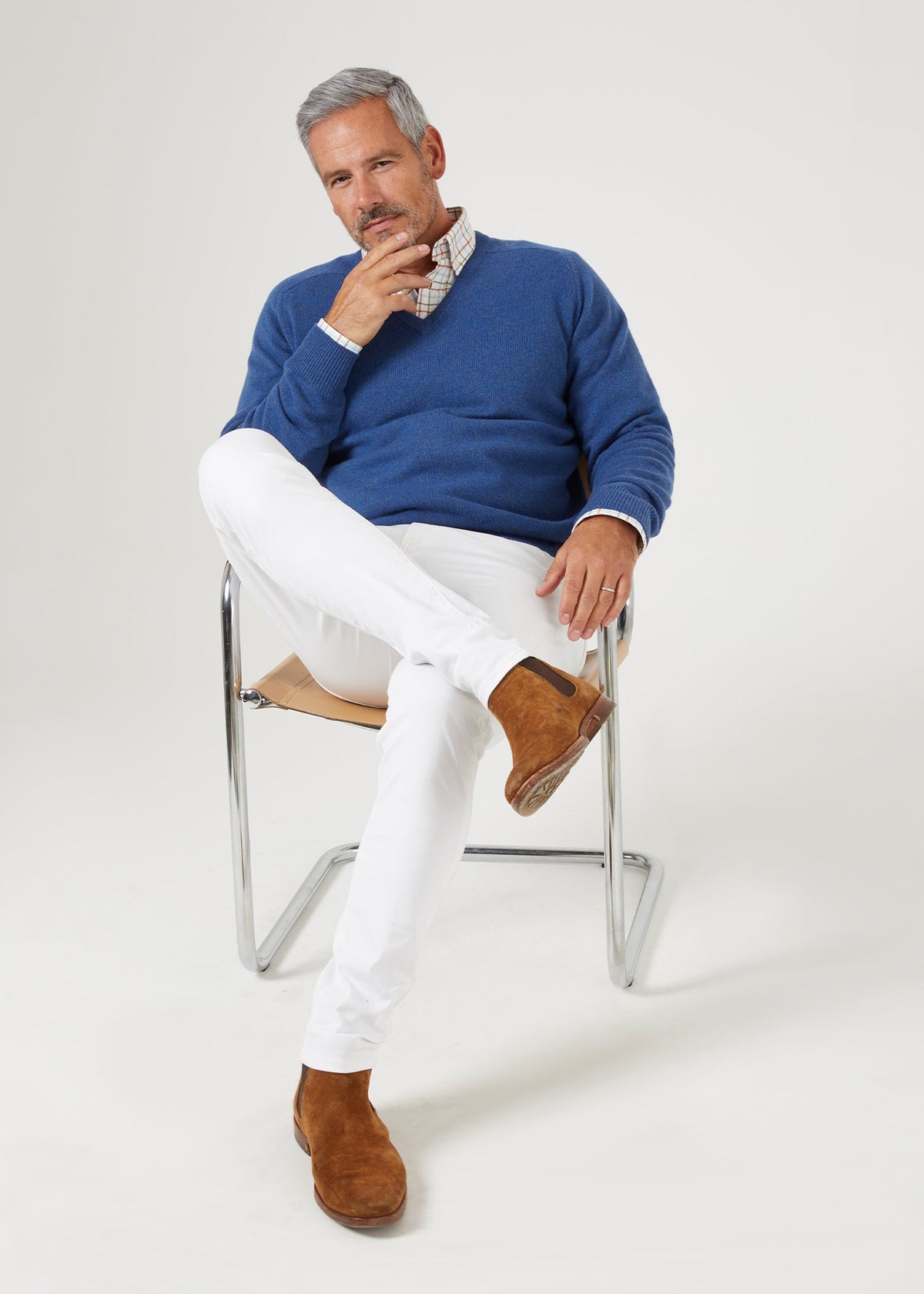 A man wearing a Selkirk Pure Cashmere V-Neck Jumper In Denim over a plaid shirt and white trousers, sitting on a chair.