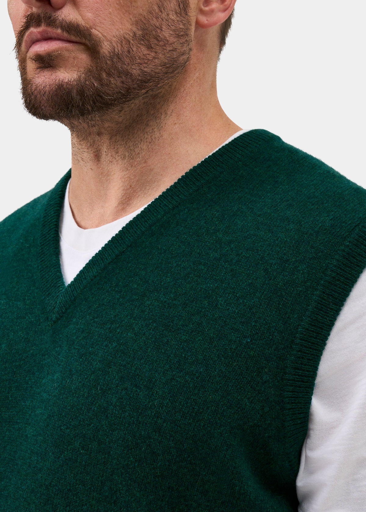A close-up of a man wearing a dark forest green knit V-neck sweater vest over a white crew-neck t-shirt, against a plain light gray background.