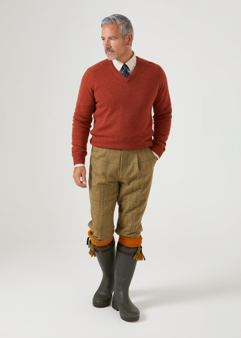 A man in a Streetly Lambswool V-Neck Jumper In Tiger, checked shirt, tie, tweed breeks, and green rubber boots.
