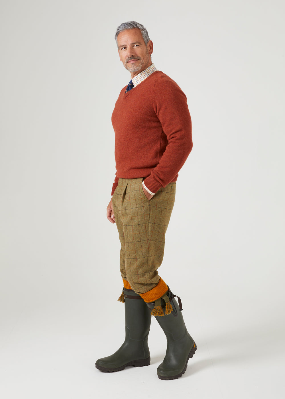 A man in a Streetly Lambswool V-Neck Jumper In Tiger over a plaid shirt and tie, with tan trousers and green rubber boots.