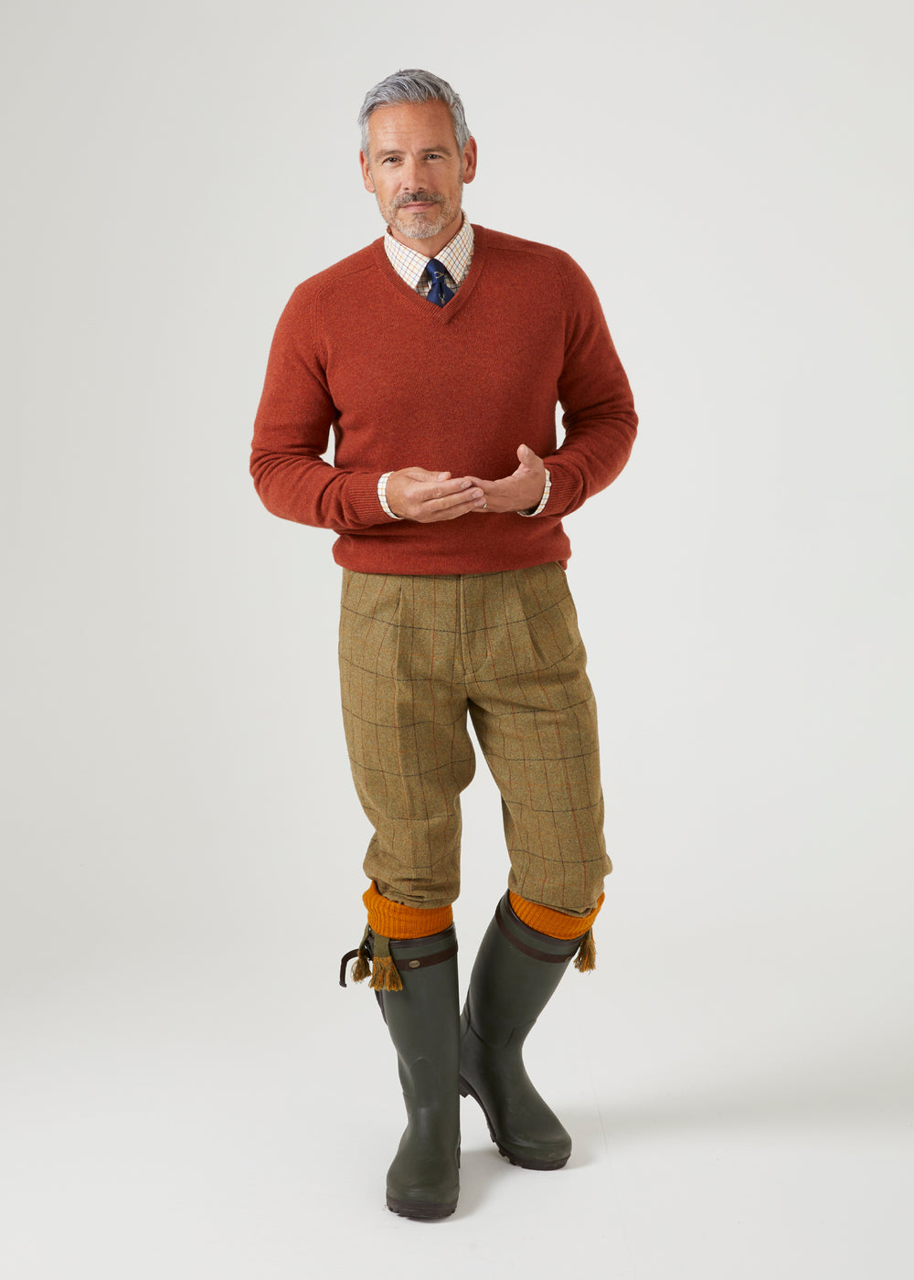 Man in a Streetly Lambswool V-Neck Jumper In Tiger, collared shirt with a tie, plaid trousers, and tall rubber boots.